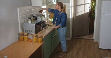 Gündelik giysiler içindeki genç bir kadın kahve çekirdeklerini parlak ve modern bir mutfakta kahve makinesine döküyor. Günlük rutin, yaşam tarzı.