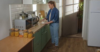 Gündelik giysiler içinde genç bir kadın parlak, modern bir mutfakta çay kaşığıyla şeker karıştırıyor. Günlük rutin, yaşam tarzı.