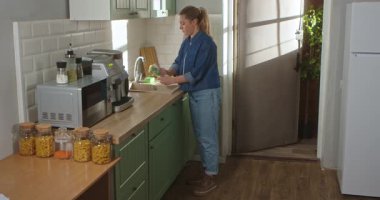 Gündelik giysiler içinde genç bir kadın parlak ve modern bir mutfakta köpüklü bir süngerle bardağı yıkıyor. Günlük rutin, yaşam tarzı.