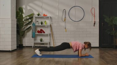 Atletik kıyafetli bir kadın, çeşitli teçhizatlarla donatılmış bir spor salonundaki paspasın üzerinde tahta egzersizi yapıyor. Egzersiz, kadınlar için fitness eğitimi, sağlıklı yaşam tarzı..