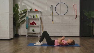 Spor kıyafetli bir kadın spor salonunda çeşitli ekipmanlarla mavi bir minder üzerinde egzersiz yapıyor. Kadın karın kasları için bir egzersiz yapıyor. Egzersiz, kadınlar için fitness eğitimi, sağlıklı yaşam tarzı..