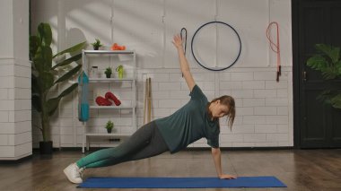 Yeşil gömlekli bir kadın modern bir fitness stüdyosunda mavi bir paspas üzerinde yan duruş sergiliyor..