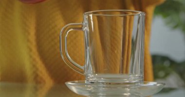 Sarı kazaklı bir kadının cam çaydanlıktan yansıtıcı bir masanın üstündeki temiz bir fincana çay dökerkenki yakın çekimi. Sıcak, rahat bir atmosfer..