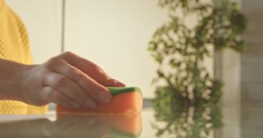 Yansıtıcı bir masayı sıcak, güneşli bir odada turuncu bir süngerle temizleyen bir el..