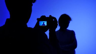 Profesyonel bir stüdyonun kulisinde bir fotoğrafçının silueti mavi ışıklı arka planda bir fotoğraf makinesiyle bir mankeni çekiyor..