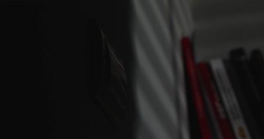 Karanlıkta bir insan eli duvardaki ışık düğmesini açar. Parlak bir ışık gelir ve beyaz duvarlı bir odayı aydınlatır ve arka planda kitaplarla dolu bir rafı..