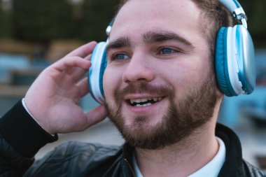 Açık havada müzik dinleyen mavi kulaklıklı, gülümseyen ve rahatlamış bir adam..