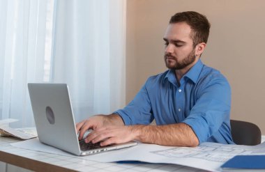 Mavi gömlekli genç adam, mimari planlarla dolu bir masada dizüstü bilgisayara odaklanmıştı..