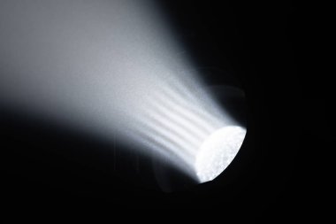 Karanlık bir odadaki spot ışıklarından yayılan güçlü, odaklanmış bir beyaz ışık demeti çarpıcı bir kontrast ve atmosfer yaratır. Sahne arkasında ışıklandırma ekipmanları.