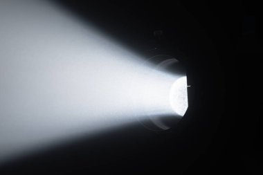 Karanlık bir odadaki spot ışıklarından yayılan güçlü, odaklanmış bir beyaz ışık demeti çarpıcı bir kontrast ve atmosfer yaratır. Sahne arkasında ışıklandırma ekipmanları.