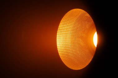 Stüdyonun spot ışığını kapat. Sıcak turuncu ışık yayıyor. Etrafını saran karanlık bir alanda dokulu bir yansıtıcı var. Sahne arkasında ışıklandırma ekipmanları.