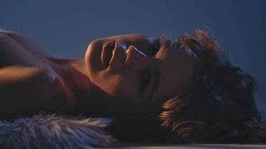 Yumuşak ışıklandırmayla aydınlatılmış, kürklü mavi bir zemin üzerinde uzanmış bir kadının yakın plan portresi..