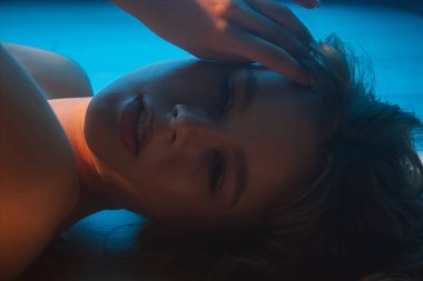 Bir kadının sakin ve sanatsal portresi uzanmış, yumuşak mavi ışıkla aydınlatılmış, sakin bir atmosfer yaratıyor..