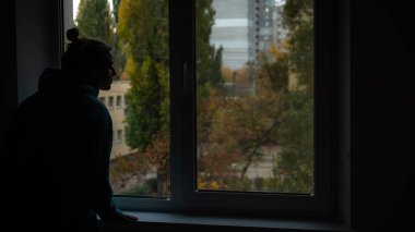 Bir bireyin silueti, arka planda ağaçlar ve binalar olan bir şehir manzarasına bakan bir pencerenin yanında duruyor..
