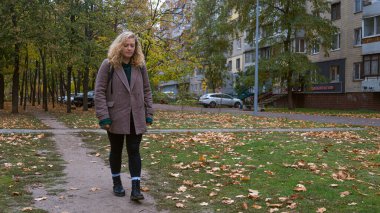 Bir kadın, ağaçlar ve konut binalarıyla çevrili bir sonbahar parkında yol boyunca yürüyor..