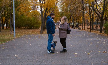 Gülümseyen bir erkek ve kadın resimli bir sonbahar parkında yan yana dururken sohbet ediyorlar..