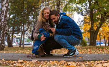 Şehrin kaldırımında diz çöküp köpekleriyle etkileşime giren neşeli bir çift. Sonbahar yaprakları onları sarıp sıcak ve rahat bir atmosfer yaratıyor..