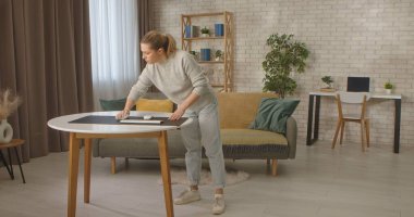 Bir kadın modern, iyi dekore edilmiş bir oturma odasında arka planda bitkiler ve masa olan bir masanın üzerine bir bilgisayar paspası siler..