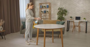 Modern dekorasyon ve ahşap mobilyalarla şık bir oturma odasında yuvarlak masa temizleyen bir kadın görülüyor..