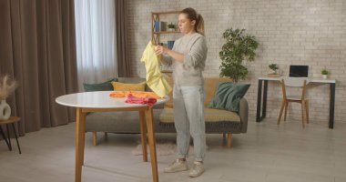 Bir kadın şık bir oturma odasında, modern ve rahat bir ortamda renkli giysileri bir masanın üzerine katlıyor..