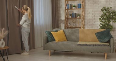 Bir kadın şık bir oturma odasında hoş gri bir kanepe ve renkli yastıklarla perdeleri temizlemek için pamuk rulo kullanır..