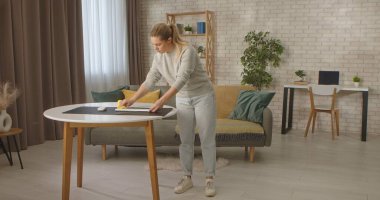 Günlük kıyafetli bir kadın modern dekorlu şık, aydınlık bir oturma odasında kablosuz bir klavyenin tozunu alır..