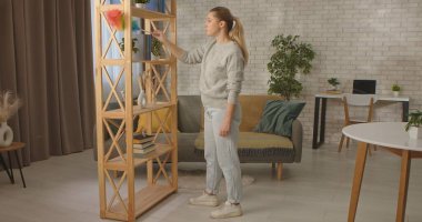 Bir kadın rahat, parlak bir oturma odasında dururken dolabın ve kitapların tozunu alıyor. Temizlik ve temizlik kavramı.