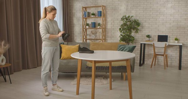 A woman stands in a stylish room, brushing a black sneaker on a white table. The room is furnished with modern furniture and filled with light.