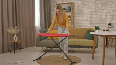 Günlük kıyafetli bir kadın, modern dekorasyonla dekore edilmiş bir oturma odasına ütü masası kuruyor..