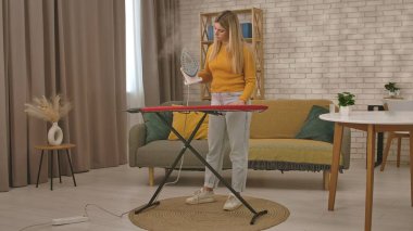 Bir kadın ütü masasının yanında elinde ütüyle duruyor ve sıcak bir evin içinde giysilerini ütülemeye hazırlanıyor..