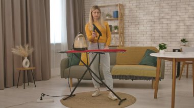 Bir kadın, modern, rahat bir oturma odasındaki ütü masasına el yapımı bir buharlı gemi kullanarak giysilerini ütülüyor..