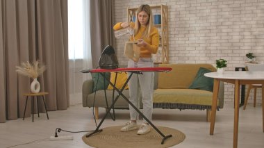 Bir kadın, modern, rahat bir oturma odasındaki ütü masasına el yapımı bir buharlı tencereye su döker..