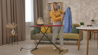 Evdeki mavi gömleği ütülemek için buhar ütüsü kullanan bir kadın, rahat bir oturma odasında ütü masasının yanında duruyor..