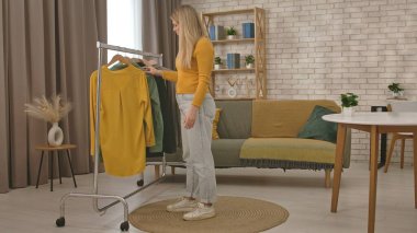 Tarafsız dekor ve doğal ışıkla modern, şık bir oturma odasından elbise seçen bir kadın..