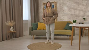 Kahverengi ceketli ve kot pantolonlu bir kadın modern iç dekorasyonlu şık bir oturma odasında duruyor..