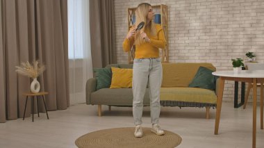 Hardal kazaklı ve kot pantolonlu bir kadın şık bir oturma odasında duruyor uzun sarı saçlarını tarıyor..