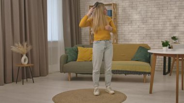 Hardal kazaklı ve kot pantolonlu bir kadın şık bir oturma odasında duruyor uzun sarı saçlarını tarıyor..
