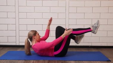 Pembe bluzlu ve siyah taytlı bir kadın mavi bir paspasın üzerinde çalışıyor ve karın egzersizleri yapıyor, spor programına odaklanıyor..