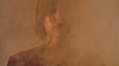 Bir kadının yakın plan portresi. Yumuşak ışıkla aydınlatılan dumandan pürüzsüz, parlak bir teni olan genç bir kadının yüzü belirir..