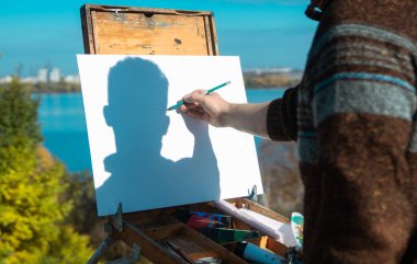 Elinde kalem tutan biri, açık bir gökyüzüne karşı boş bir tuval üzerinde resim çiziyor..