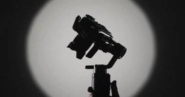 Operatör, motorlu dengeleyiciye monte edilmiş, süspansiyonlu, koyu arkaplanda parlak bir yuvarlak ışığa karşı çekilen, film yapımını sembolize eden kamerayı sabitler..