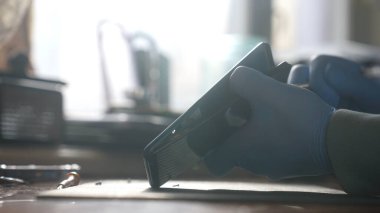 Mavi eldiven giyen bir kişi, güneşli bir atölyede masa başında çalışırken kırık bir akıllı telefon tutar..