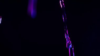 Parlayan mavi ve mor su damlacıklarının ve akıntılarının soyut yakın plan derin siyah bir arkaplan üzerine düşmesi, dramatik neon ışıkla aydınlatılması ve metin veya tasarım için merkezde boş kopya alanı bırakılması.