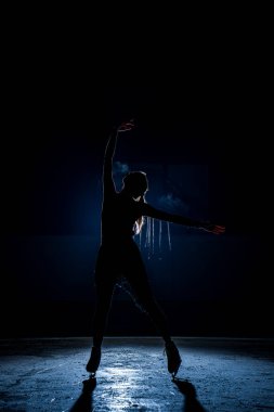 Bir artistik patinajcının silueti açık mavi arka plan ışığı ve sisli kapalı bir buz pateni pistinde performans sergiliyor. Şık duruşu, hareketi ve tiyatral atmosferi olan dramatik kış sporları sahnesi.