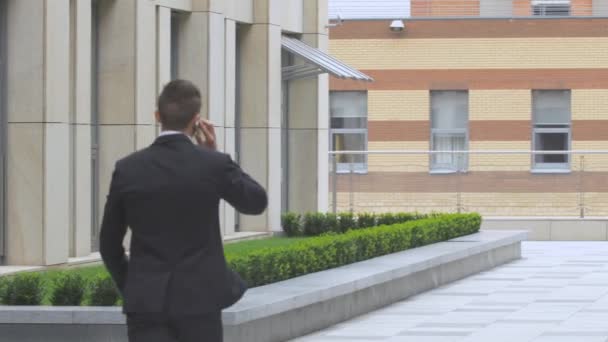 Young man speaking on the phone while walking along modern building — Αρχείο Βίντεο