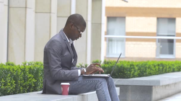 Hombre de negocios americano que trabaja con gafas en el ordenador portátil al aire libre en un fondo de edificio corporativo — Vídeo de stock