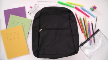 Aerial stop-motion shot of a school bag being packed with colourful supplies and a traditionally female toy, on a plain background.