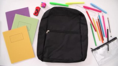 Aerial stop-motion shot of a school bag being packed with colourful supplies and a traditionally male toy, on a plain background.