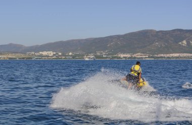 Tatil sırasında bir turist, sıcak bir yaz gününde sıcak bir jet ski zirvesinde koruyucu bir kıyafetle dağların arka planına doğru koşar. resim bulanıklaştırma