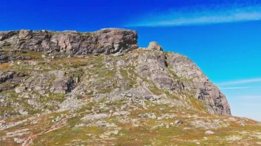 Büyük, tıkalı bir kayanın alçak açılı görüntüsü ve dik bir dağ yamacı Norveç 'teki canlı, derin mavi gökyüzünün altındaki engebeli dağlık araziyi gözler önüne seriyor..